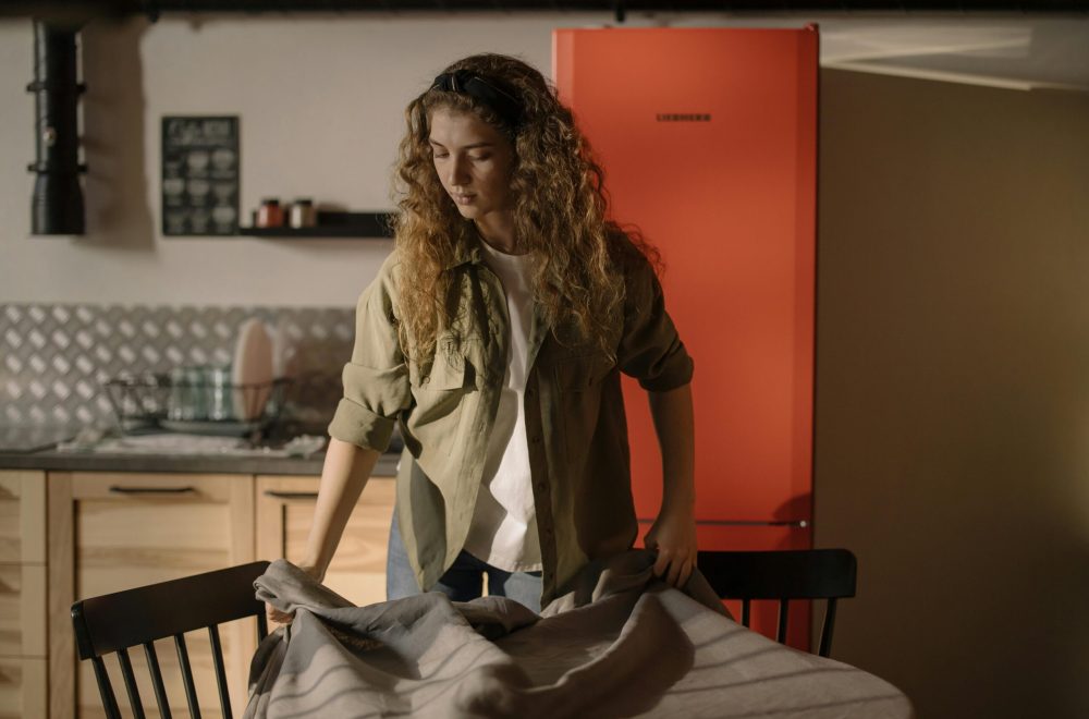 A woman in a casual kitchen setting a dinner table with a tablecloth.